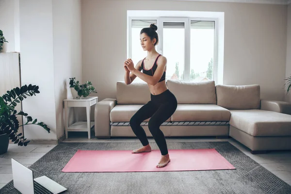 Brunette girl doing warming exercises at home on a pink sport carpet with a laptop — Stock Photo, Image