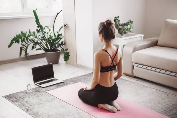 Mujer caucásica sentada frente al portátil y practicando yoga en casa vestida con ropa deportiva sobre una alfombra deportiva rosa — Foto de Stock