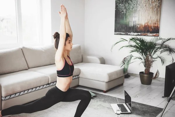 Jovencita con el pelo negro estirando su cuerpo mientras hace yoga delante de la computadora usando ropa deportiva — Foto de Stock
