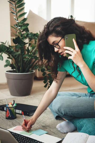 Занятая белая женщина с вьющимися волосами делает заметки, сидя за компьютером и разговаривая по телефону — стоковое фото