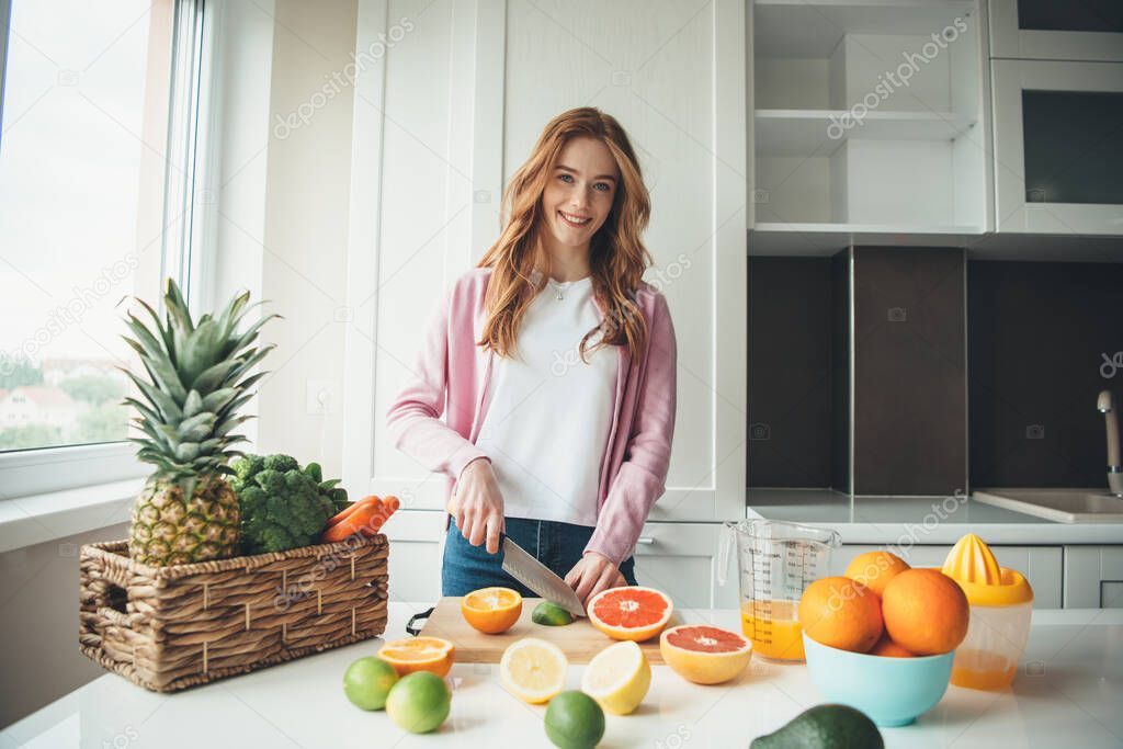 Caucasian woman with red hair and freckles smiling at camera while cutting fruits and squeezing them making juice from lime and orange