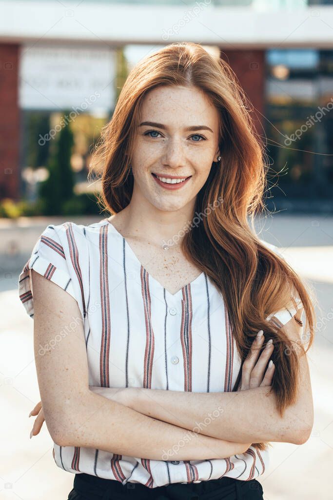 Gorgeous caucasian woman with freckles and red hair posing with crossed hands and smile at camera outside
