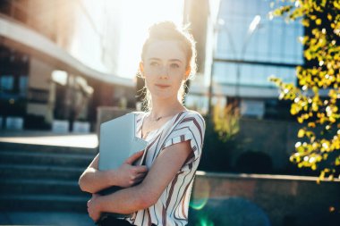Güneşli bir günde dizüstü bilgisayar ve cep telefonunu kucaklayan kızıl saçlı ve çilli bir kadının ön görüntüsü.