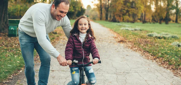 Dikkatli bir baba kızına bisiklet sürmeyi öğretirken ona parkta gülümserken yardım ederken — Stok fotoğraf
