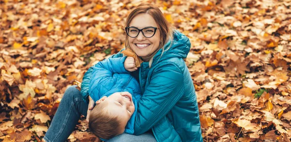 Mavi gözlüklü sarışın beyaz anne ve oğlu yerde yapraklarla oynuyorlar. — Stok fotoğraf