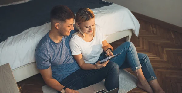 Upper view photo of a caucasian couple lying on the floor in the bedroom and smiling cheerfully while using a tablet
