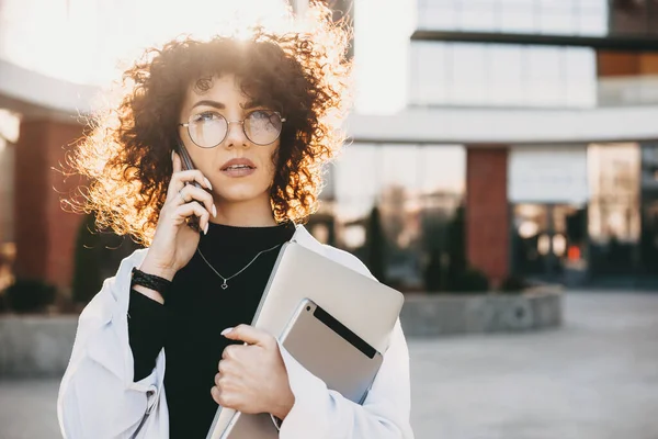 Ung entreprenör med lockigt hår och glasögon talar i telefon medan poserar med en bärbar dator utanför — Stockfoto