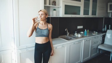 Spor kıyafetleri içinde sarışın bir kadın egzersiz yaptıktan sonra mutfakta su içiyor.