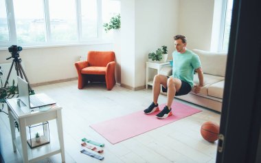 Evde spor yapan beyaz bir antrenör kamera ve dizüstü bilgisayarla online özel ders veriyor.