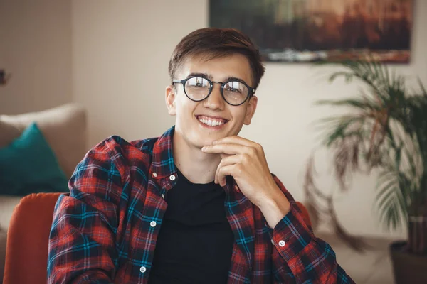 Jovem caucasiano com óculos vestindo uma camisa está posando na poltrona tocando seu queixo e sorrir para a câmera — Fotografia de Stock
