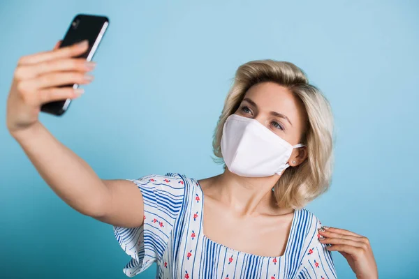 Bella signora bionda con maschera medica sul viso sta facendo un selfie utilizzando un telefono su un muro di studio blu — Foto Stock