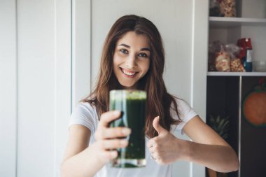 Kahverengi saçlı çekici beyaz kadın kameraya gülümserken işaret gibi el kol hareketi yapıyor ve elinde taze sebze suyuyla bir bardak tutuyor.