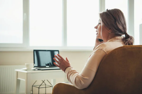 Senior caucasian businesswoman talking on phone while sitting in armchair with a laptop in front — Stock Photo, Image