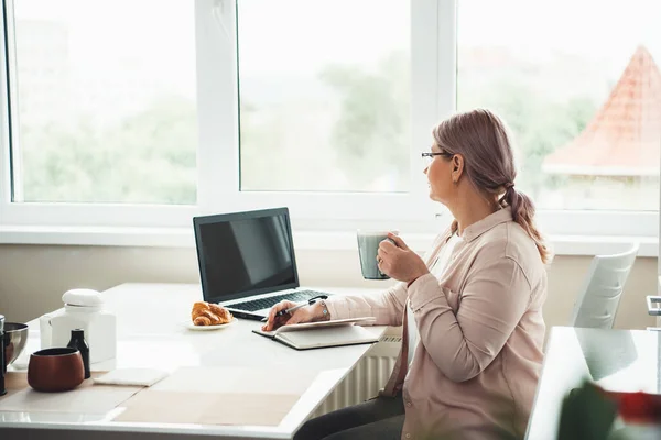 Senior woman working from home during the quarantine looking at the window while drinking tea with croissant and work at computer — Stock Photo, Image