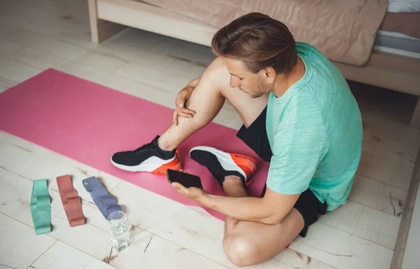 Hombre caucásico con cabello rubio usando ropa deportiva está buscando en el teléfono para clases de acondicionamiento físico en casa antes de empezar a hacer ejercicio — Foto de Stock