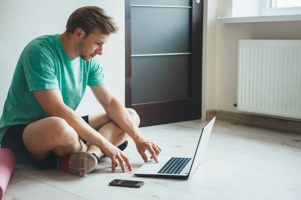 Uomo caucasico con barba e capelli biondi seduto sul pavimento e utilizzando un computer portatile prima di praticare yoga con un tappeto speciale — Foto Stock