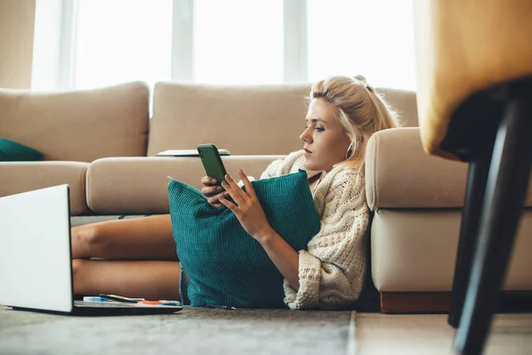 Mujer encantadora con el pelo rubio charlando en el móvil mientras está sentado en el suelo y haciendo la tarea —  Fotos de Stock