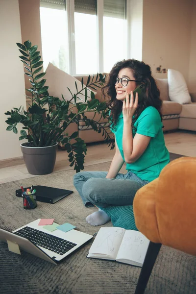 Милая кавказская девушка в очках и кудрявые волосы разговаривают по телефону, сидя на полу и имея онлайн-уроки — стоковое фото