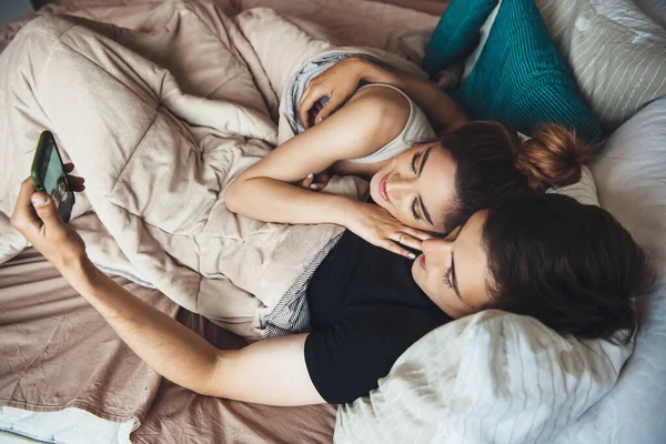 Casal jovem deitado na cama e abraçando enquanto sorri e fazendo selfie coberto com uma colcha de manhã cedo — Fotografia de Stock