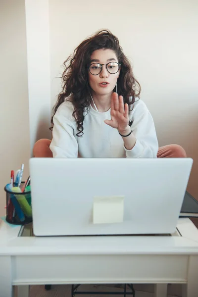 Young caucasian girl with curly hair and eyeglasses is having online lessons at the university sitting at the laptop with earphones Royalty Free Stock Photos