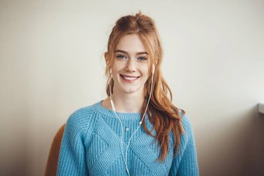 Kafkasyalı, kızıl saçlı, çilli, koltukta oturan ve kulaklıkla müzik dinlerken kameraya gülümseyen bir kadının ön görüntüsü.