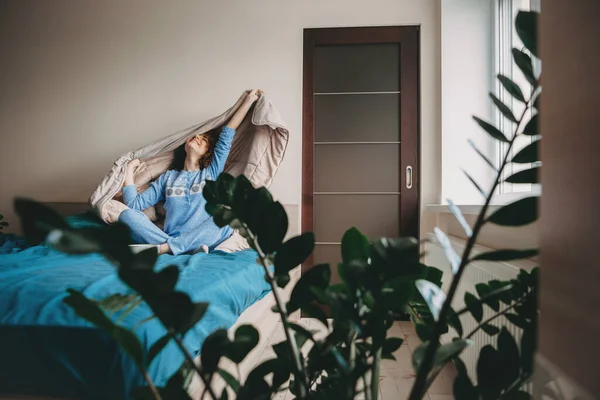 Dame caucasienne se réveille de son lit habillé d'un pyjama bleu souriant et étirant avec sa courtepointe — Photo