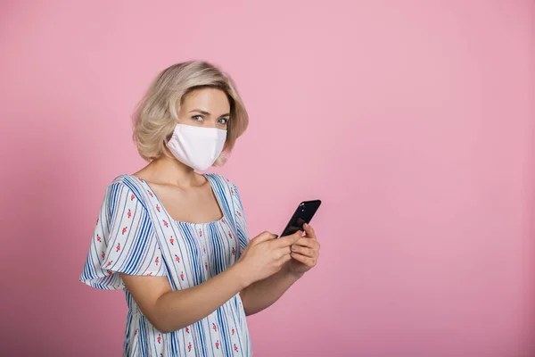 Femeia fermecătoare cu părul blond purtând o mască medicală vorbește la telefon și se uită la camera de pe un perete de studio roz cu spațiu liber — Fotografie, imagine de stoc