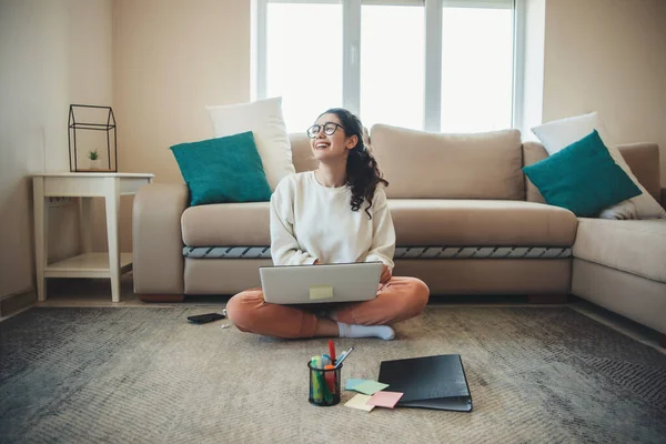 Busy university girl wearing eyeglasses and curly hair is studying on floor with a laptop during online courses Stock Picture