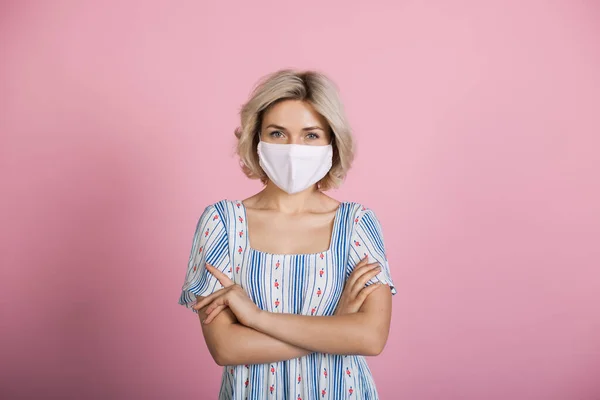 Blonde kaukasische Frau mit medizinischer Maske und Kleid blickt an einer rosa Studiowand in die Kamera — Stockfoto