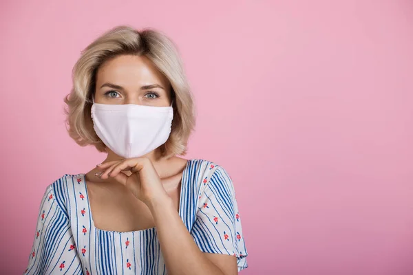 Blondine vrouw met een medisch masker op het gezicht kijken naar de camera tijdens het dragen van een jurk en het aanraken van haar kin op een roze studio muur met vrije ruimte — Stockfoto