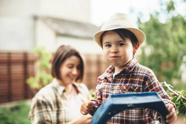 Affascinante ragazzo caucasico con un cappello in testa mangiare ciliegie dall'albero in posa con la madre sullo sfondo sorridente — Foto Stock