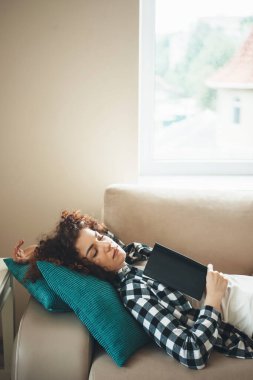 Kitap okuduktan sonra kanepede dinlenen kıvırcık saçlı uykulu beyaz kadın.
