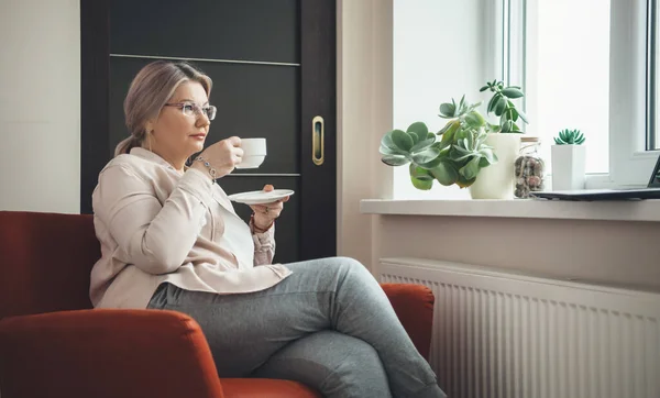 Senior caucasian woman with eyeglasses sitting in armchair and drinking a coffee while wearing eyeglasses and watching a lesson on laptop — Stock Photo, Image