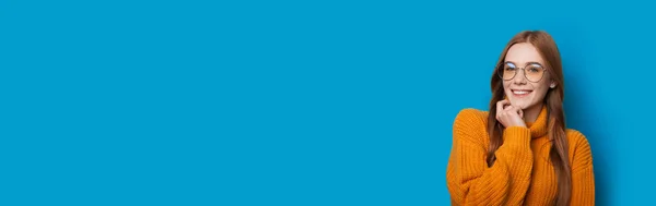 Ingwer junge kaukasische Dame mit Sommersprossen und Brille berührt ihr Kinn und lächelt auf einer blauen Studiowand mit freiem Platz — Stockfoto