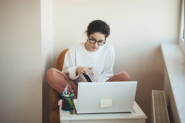 Vit student med glasögon sitter i fåtölj gör läxor med hjälp av en bok och bärbar dator — Stockfoto