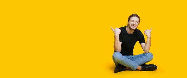 Homem caucasiano com barba e cabelos longos sentado no chão e gesticulando o sinal semelhante perto do estúdio amarelo espaço livre — Fotografia de Stock