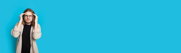 Homem caucasiano cuidadoso com cabelos longos e óculos está tocando sua cabeça e olhando para a câmera em uma parede de estúdio azul com espaço livre — Fotografia de Stock