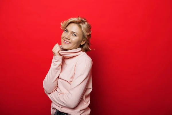 Bella donna caucasica con i capelli biondi con un maglione rosa sorride alla telecamera su un muro rosso studio — Foto Stock