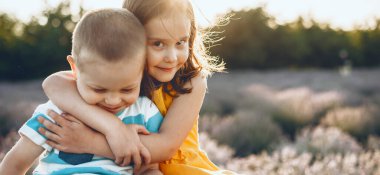 Beyaz erkek ve kız kardeşler kameraya bakıp gülümserken lavanta tarlasında poz veriyorlar ve kucaklaşıyorlar.
