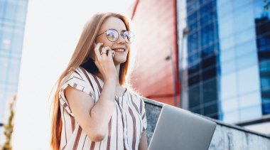 Kırmızı saçlı, gözlüklü ve çilli bir girişimci şehrin dışında oturuyor ve telefonda konuşurken dizüstü bilgisayarın başında çalışıyor.