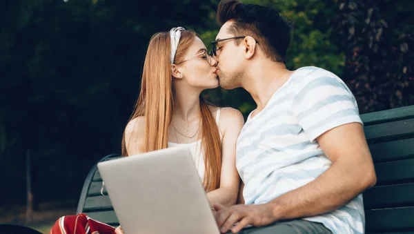 Härligt par sitter på bänken och kysser när du använder en bärbar dator på en solig dag — Stockfoto