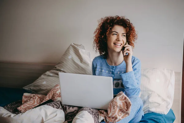 Joven dama caucásica con el pelo rizado hablando por teléfono mientras sonríe y sostiene un ordenador portátil en la cama — Foto de Stock