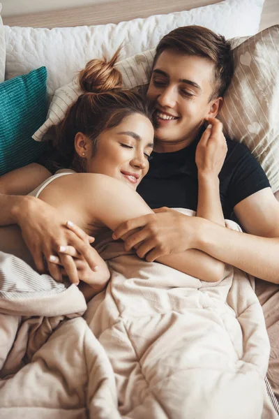 迷人的高加索夫妇躺在床上，清晨拥抱着他们，闭着眼睛笑着 — 图库照片