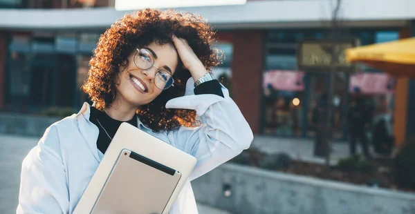 Mulher encantadora com cabelo encaracolado posando fora com um laptop tocando seu corte de cabelo enquanto sorri para a câmera — Fotografia de Stock