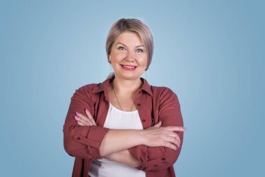 Mavi bir stüdyonun duvarındaki kameraya gülümseyen çapraz elli çekici yaşlı kadın.