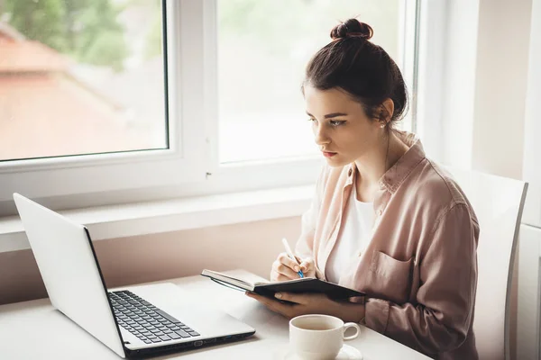 Концентрированная белая женщина в рубашке пишет рецепт в книге, глядя на ноутбук и выпивая чашку чая у окна — стоковое фото