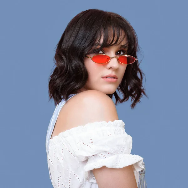 Preciosa mujer morena con gafas naranjas mirando hacia atrás a la cámara y posando en una pared de estudio azul — Foto de Stock