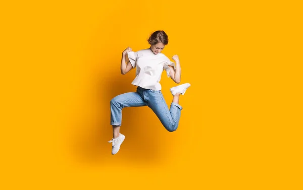 Saltando mulher caucasiana em jeans e camiseta branca sorrindo e anunciando algo em uma parede de estúdio amarelo — Fotografia de Stock