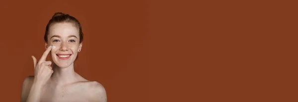 Рыжая белая женщина с обнаженными плечами и веснушками, рекламирующая крем на коричневой стене студии, улыбаясь и нанося его на лицо — стоковое фото
