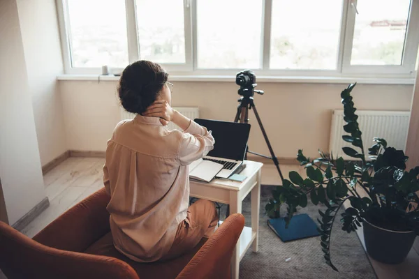 Mujer caucásica cansada en ropa formal que tiene reunión en línea con una cámara y un ordenador portátil mientras está sentada en la mesa en un sillón — Foto de Stock